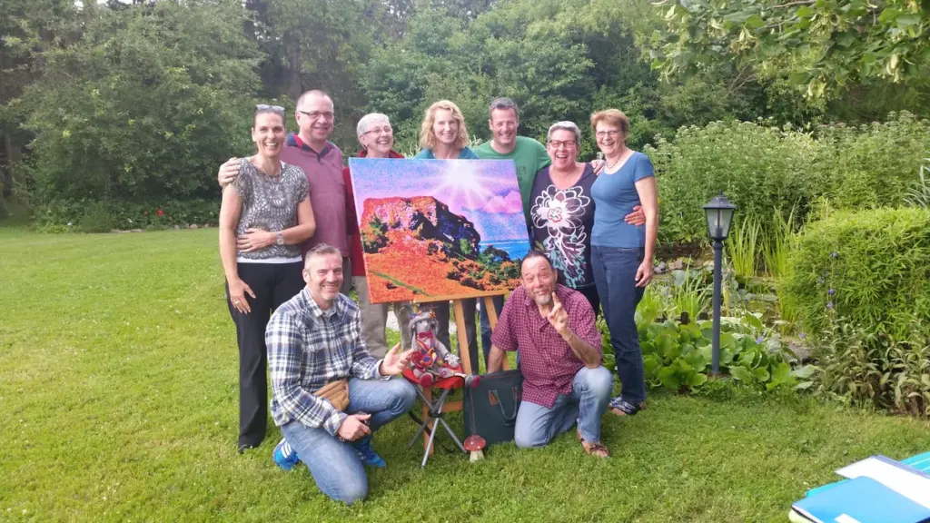 Neun Personen stehen und knien in einem Garten um ein farbenfrohes Landschaftsbild auf einer Staffelei.