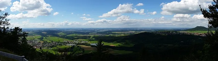 Panoramablick über grüne Felder, Ortschaften und bewaldete Hügel unter blauem Himmel mit weißen Wolken.