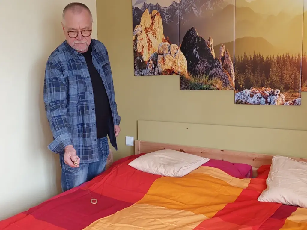Ein Mann hält ein radiästhetisches Instrument über ein Bett.
